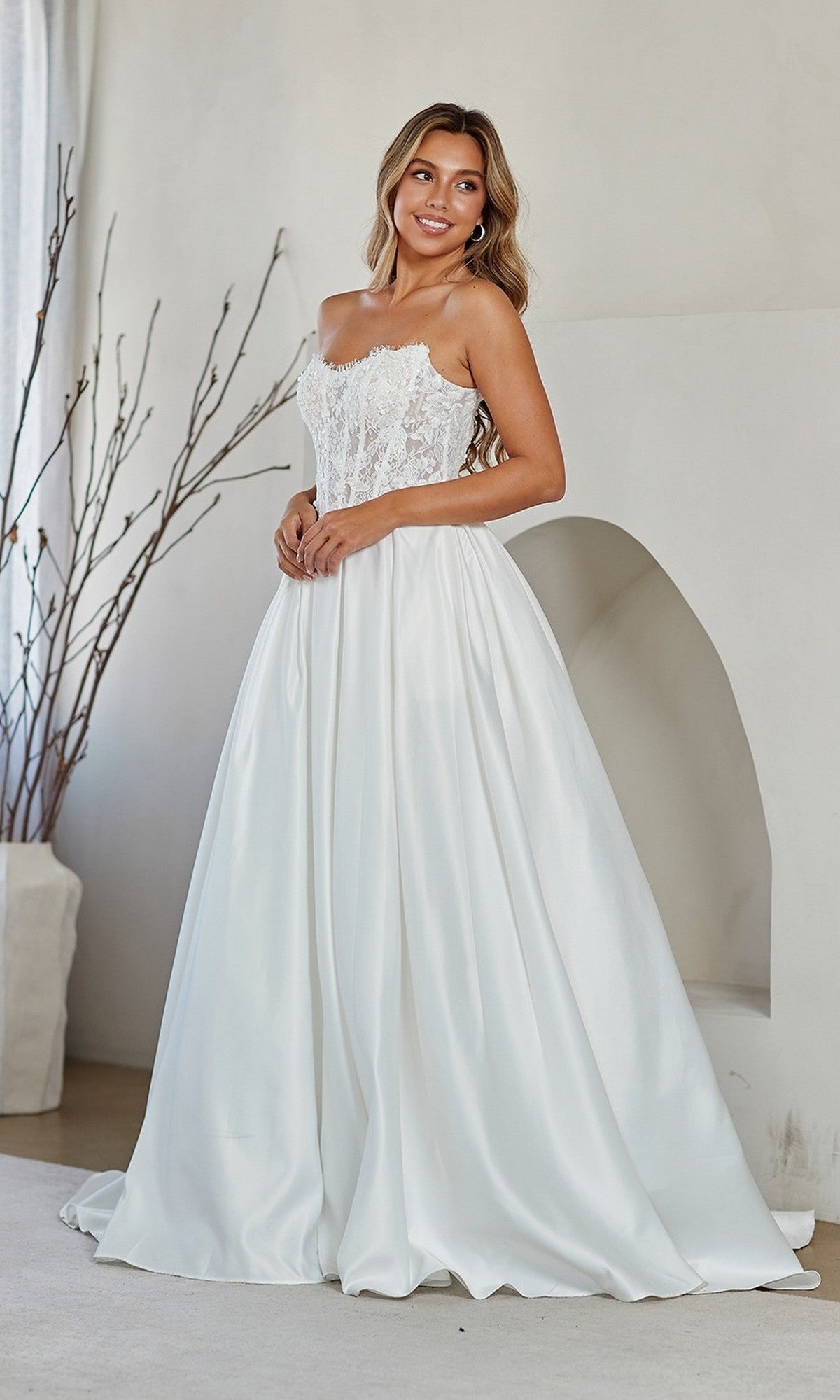 A woman wearing the Amelia Couture 2089 long prom dress with a strapless white lace corset bodice and satin skirt stands indoors near a minimalist archway and decorative branches.